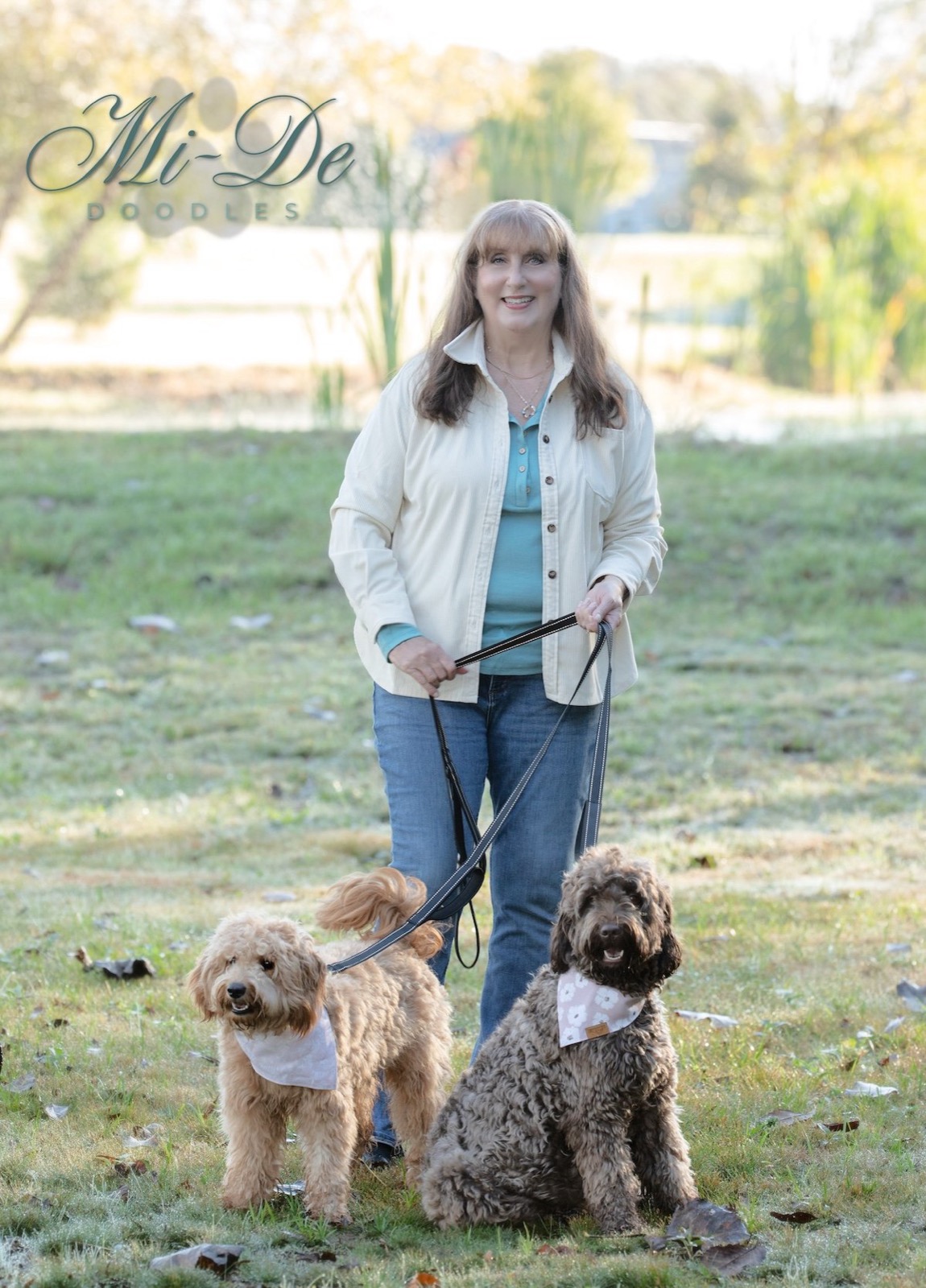 A smiling woman in a light jacket and jeans stands outdoors on grass, holding leashes attached to two fluffy goldendoodles wearing bandanas. The background features trees, a pond, and sunlight.