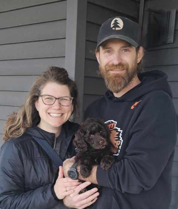 A smiling couple stands in front of a dark gray house, both wearing glasses and jackets. The man holds a small brown puppy, while the woman rests her hand on the puppy. All three look at the camera.