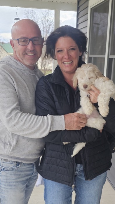 A smiling man and woman stand close together outside, with the woman holding a fluffy light-colored puppy. They both wear casual clothes, and the background shows a house and trees.
