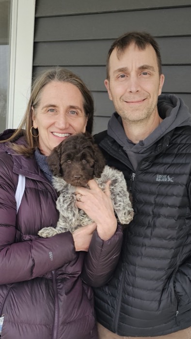 Smiling couple holds a brown curly puppy in front of a dark siding house wall outside.