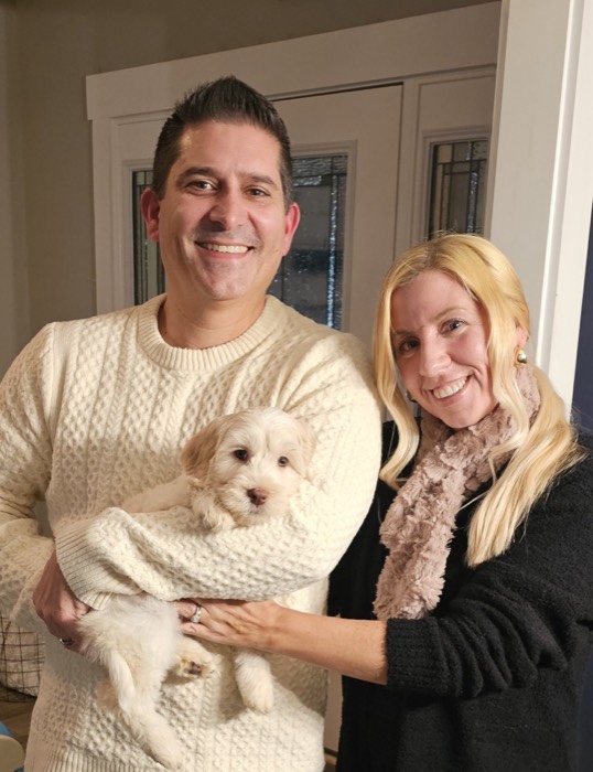 A smiling man in a cream sweater holds a small, fluffy puppy, while a woman with long blonde hair and a scarf stands beside him, both posing happily indoors.