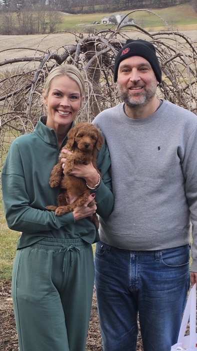 A smiling woman in a green tracksuit holds a small brown puppy, standing next to a smiling man in a gray sweatshirt and black beanie. They are outdoors with leafless trees and a rural landscape in the background.