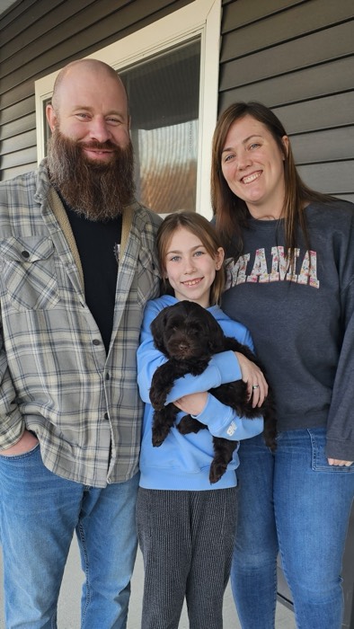A bearded man, a woman, and a smiling girl holding a small black puppy stand together and pose for a photo in front of a dark house with a window.