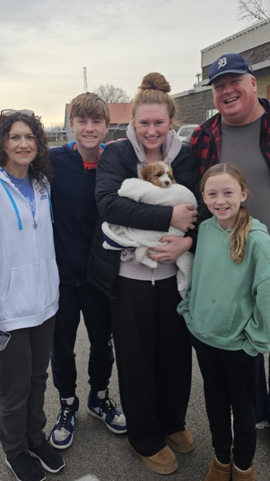 Group of five smiling people posing outdoors, a young woman cradling a small brown puppy wrapped in a white blanket.