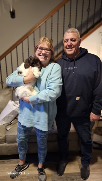 A smiling woman in blue holds a fluffy white and brown puppy, standing next to a man in a dark hoodie. They pose indoors in front of a couch and staircase.