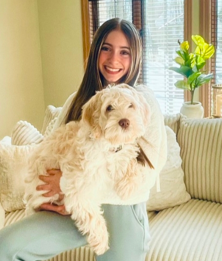 A smiling young woman with long brown hair sits on a light-colored couch, holding a fluffy cream-colored dog. Sunlight streams in through the window behind them, and a green leafy plant is on a side table.
