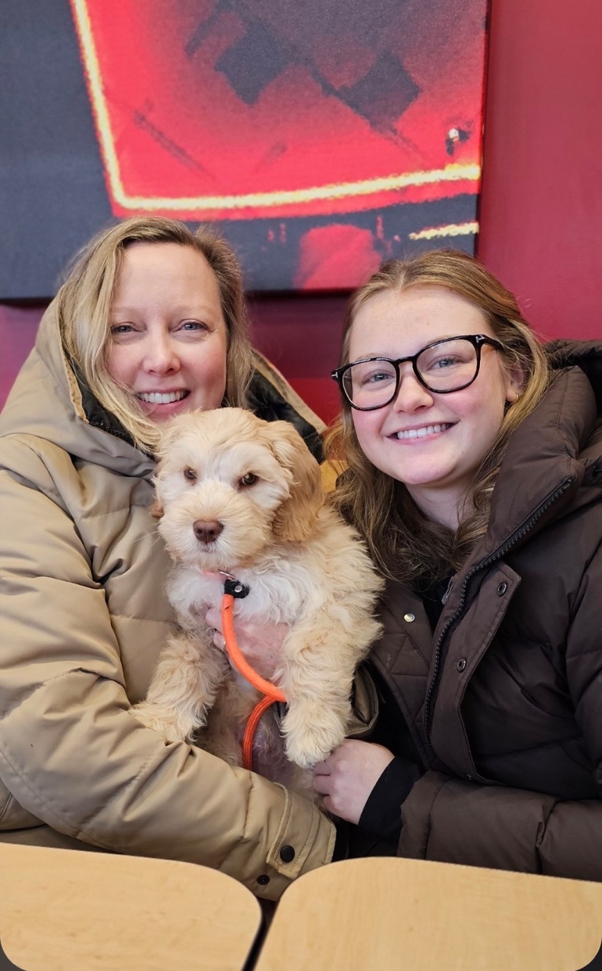 Two women wearing winter coats are smiling and sitting closely together, holding a fluffy light-colored puppy with an orange leash, against a red and black background.