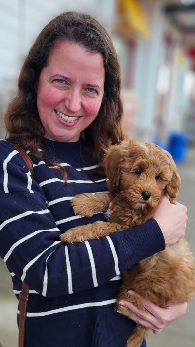 A woman with wavy brown hair and a navy striped sweater smiles while holding a fluffy tan puppy in her arms outdoors.