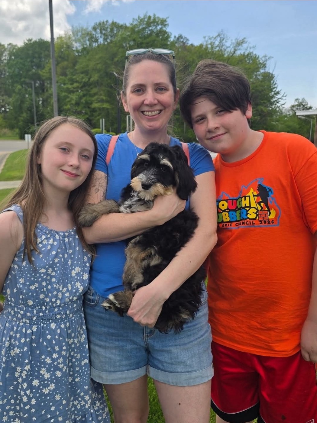 A smiling woman holds a small black and tan dog, standing outside with a girl in a blue dress and a boy in an orange shirt. Trees and grass are visible in the background.