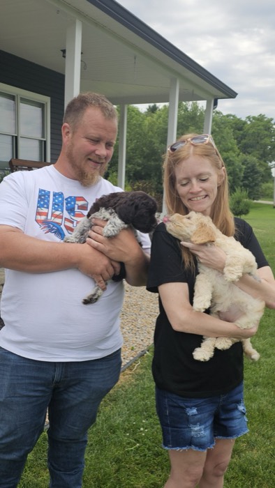 A man and woman stand outside holding two puppies. The man, in a white USA flag shirt, holds a dark puppy, while the woman, in a black shirt, holds a light puppy thats licking the other puppy. Both adults are smiling.