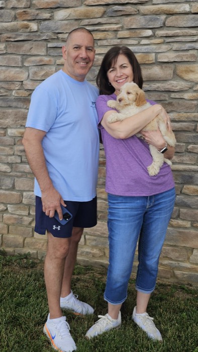 A man in a light blue shirt and dark shorts stands next to a woman in a purple shirt and jeans who is holding a small tan puppy. They are smiling in front of a stone wall, standing on grass.