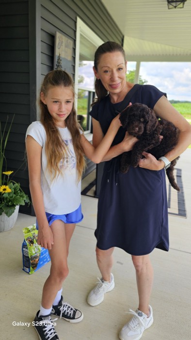 A woman in a navy dress holds a curly brown puppy while standing next to a young girl in blue shorts and a white shirt on a porch. Both are smiling; a bag of dog food is on the ground beside them.