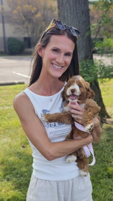 A woman in a sleeveless white top smiles while holding a small brown and white puppy outside on a sunny day, with trees and grass in the background.