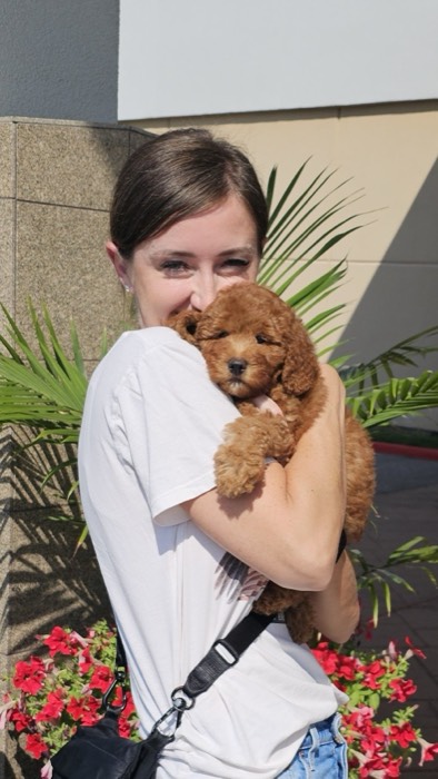 Woman holding a fluffy brown poodle puppy outdoors, smiling at the camera.