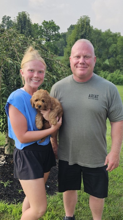 A smiling woman holds a small brown puppy next to a man. They are outdoors on green grass with trees and a bush in the background.