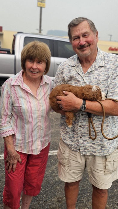 Two smiling adults stand in a parking lot; the man holds a small brown dog. They wear casual clothes and appear happy. A truck and building are visible in the background.