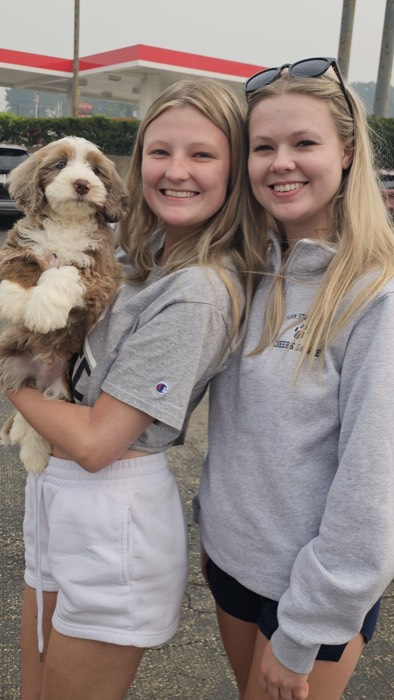 Two smiling young women stand close together outdoors. One woman holds a fluffy brown and white puppy. Both women have long blonde hair and are dressed in casual clothes. The background shows cars and part of a building.