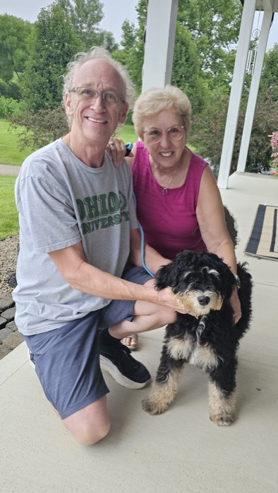 An older man and woman smile on a porch while kneeling beside a fluffy black and tan dog. The man wears an Ohio University shirt and glasses. Green trees and grass are visible in the background.