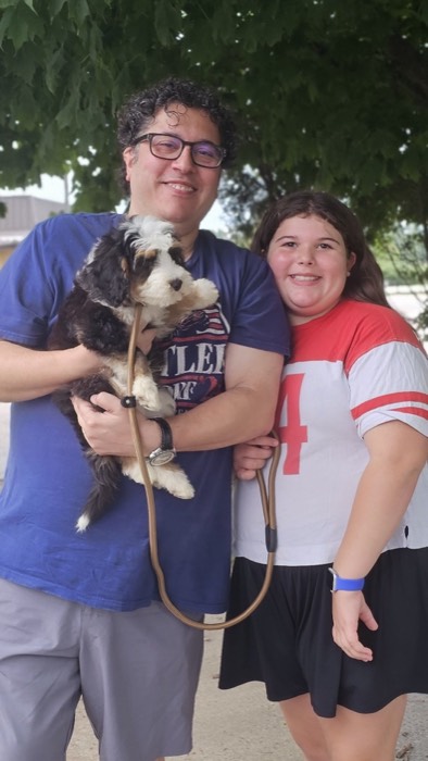 A man holding a black and white dog stands next to a girl in a red and white shirt under a tree. Both are smiling at the camera.