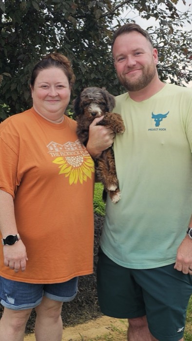 Two smiling adults standing outdoors, the woman in an orange shirt and the man in a light green shirt. They are holding a small brown and black puppy between them. Bushes and grass are visible in the background.