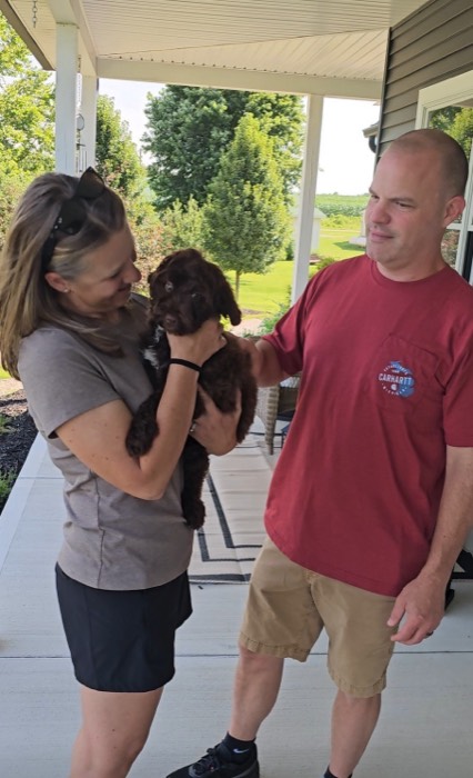 A woman holds a small brown puppy and smiles at it while a man in a red shirt stands beside her, gently petting the puppy. They are standing on a porch with green trees in the background.