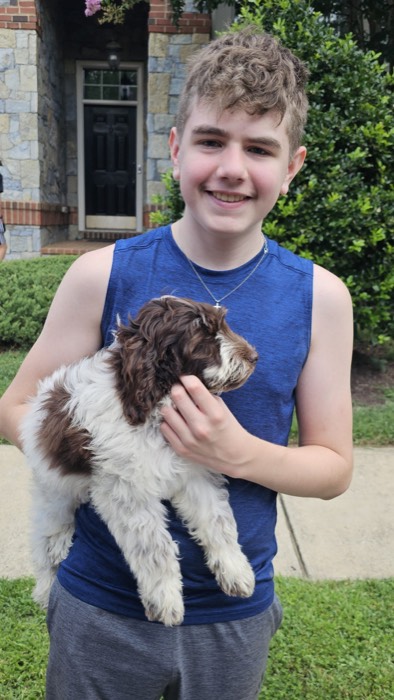 Smiling boy in a blue sleeveless shirt holds a fluffy brown-and-white puppy outdoors in front of a stone house.