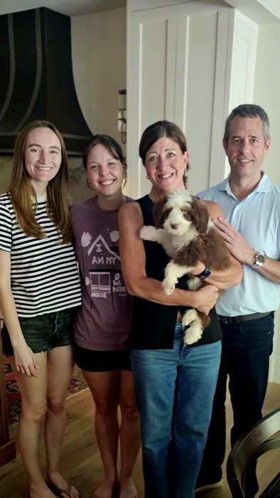 Four adults smile for a photo indoors, with a fluffy brown-and-white puppy held by the woman in a black sleeveless top in the center.