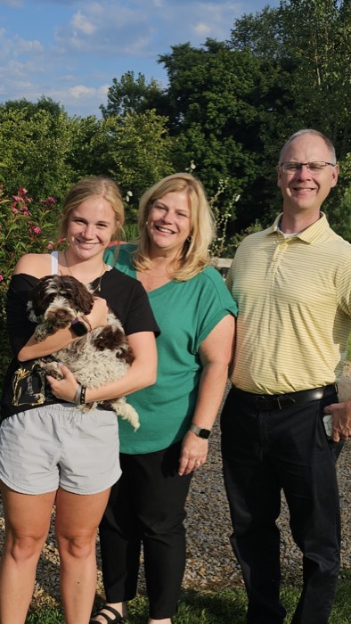 Three adults stand outside smiling with a young woman holding a small black and white dog. Trees and flowering plants are in the background. The weather appears sunny and pleasant.