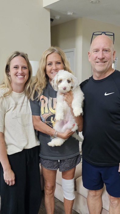 Three adults smile at the camera indoors; the woman in the center holds a fluffy white puppy. The group stands close together in a living room with neutral-colored walls.