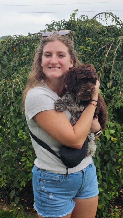 A smiling woman with long hair, wearing a gray shirt and blue shorts, stands outdoors holding a small, fluffy brown and white dog. Lush green foliage fills the background.