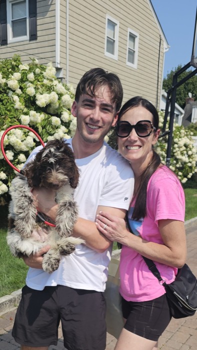A smiling man holding a fluffy brown and white dog stands next to a smiling woman in sunglasses. They are outdoors in front of a house and blooming white flowers.