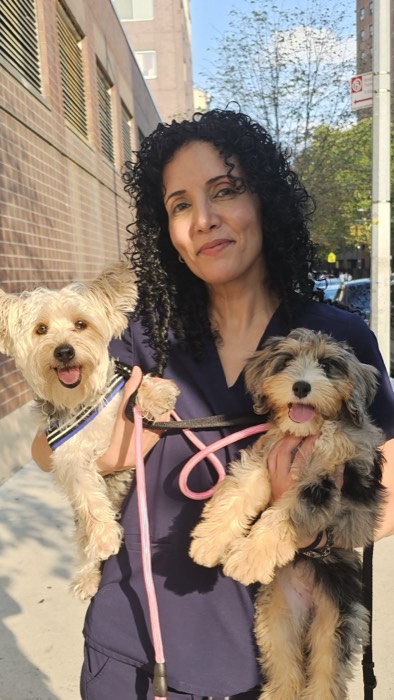 A woman with curly hair stands on a sunny city sidewalk, smiling and holding two small, fluffy dogs on leashes. Both dogs are happy with their tongues out. Trees and buildings are visible in the background.