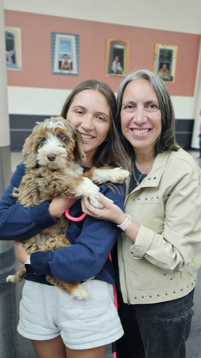 Two women smiling and holding a fluffy brown and white puppy indoors, with framed photos on a pink and beige wall behind them.