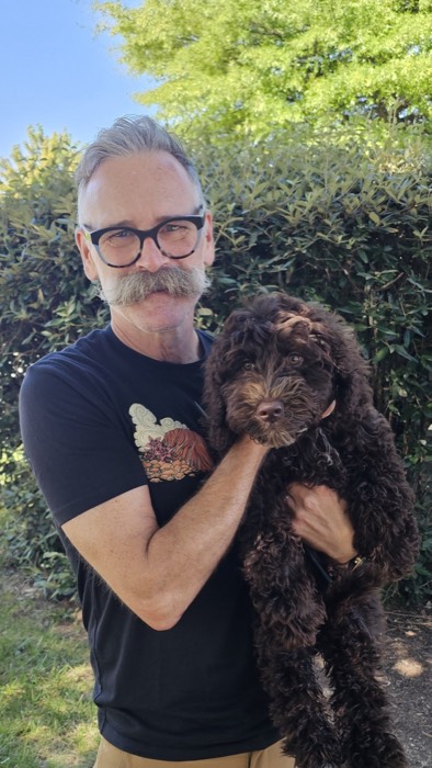 A man with glasses and a mustache holds a fluffy brown dog outside, standing in front of green bushes and trees on a sunny day.