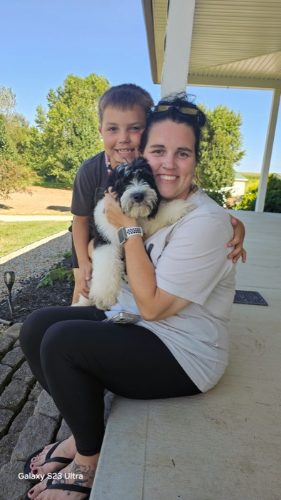 A smiling woman sits on a porch holding a fluffy black and white puppy, while a young boy hugs her from behind. Trees and a lawn are visible in the background.